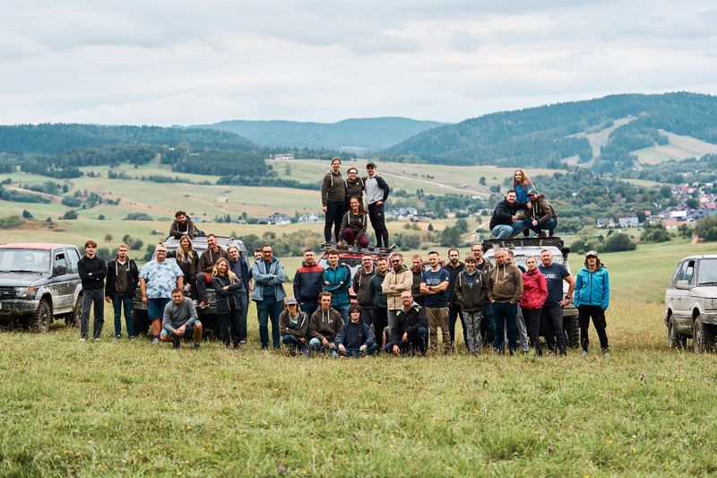 Zdjęcie grupowe uczestników integracji firmowej Novomatic podczas wyprawy off-road. Pracownicy pozują przy samochodach terenowych na tle malowniczych gór i zielonych wzgórz.