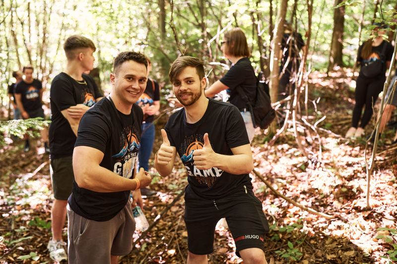 Dwóch pracowników NTP podczas ćwiczenia integracyjnego w lesie, pokazujących kciuki w górę.