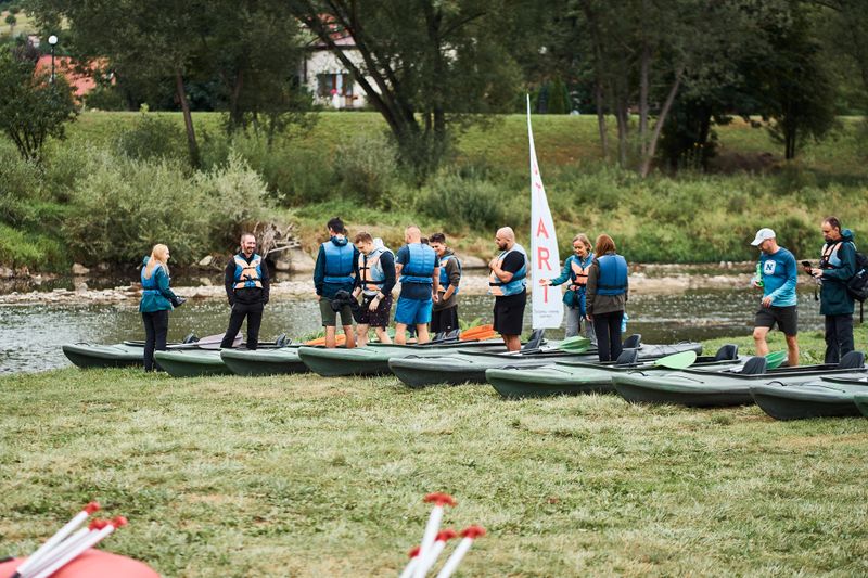 Uczestnicy integracji firmowej Novomatic przygotowują się do spływu kajakowego, stojąc w kamizelkach ratunkowych nad brzegiem rzeki, w otoczeniu zieleni.